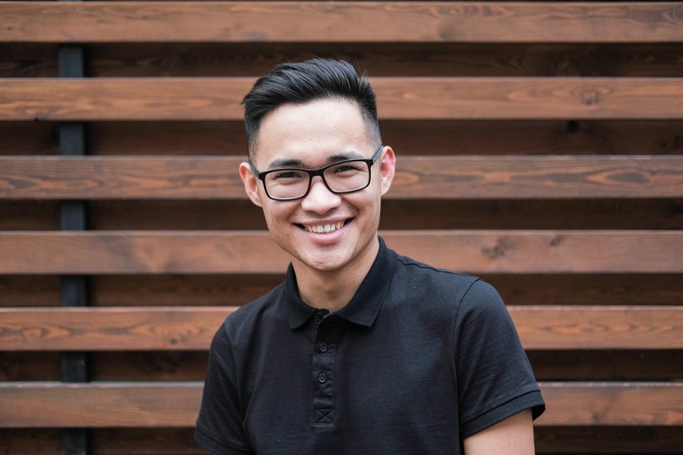 Cheerful young Asian man in eyeglasses standing against plank wall